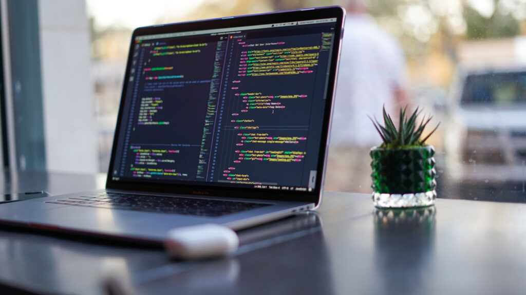 Ein moderner Arbeitsplatz mit einem Laptop auf einem Holztisch, auf dessen Bildschirm Programmiercode zu sehen ist.