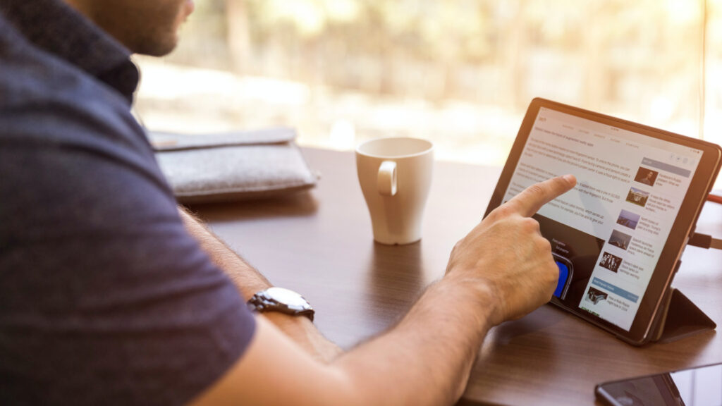 Ein Mann sitzt an einem gut organisierten Arbeitsplatz mit Tablet. Er zeigt auf eine Website.